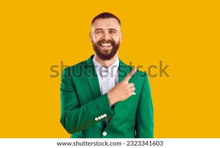 Cheerful handsome bearded man in green suit is pointing with his finger to side for advertising space. Portrait of Caucasian smiling man on orange background. St. Patrick's Day concept.