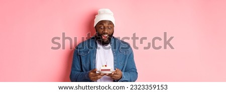 Cheerful african-american guy celebrating birthday, making wish on bday cake with lit candle, smiling happy, standing over pink background.