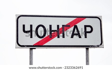 Road sign with text Chongar Chonhar, name of Ukrainian city in Kherson oblast, crossed out with red line, on white background.