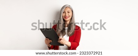Elegant asian senior businesswoman working with documents, holding pen and clipboard, signing contract and smiling, standing over white background.