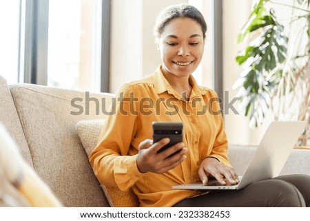 Modern smiling African American business woman, freelancer holding mobile phone, using laptop working online from home. Stylish copywriter with smartphone sitting in cafe. Successful business  