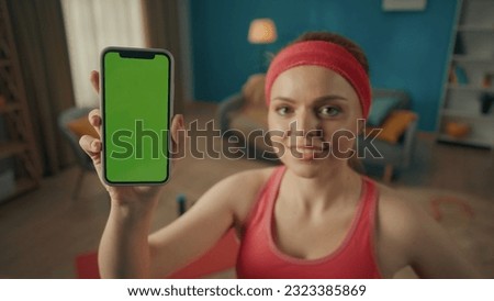 Portrait of a woman in a pink tracksuit and headband in the living room after a workout close up. A woman shows the screen of a smartphone with a green screen. Advertising area, workspace mock up.