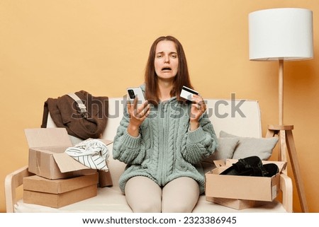 Sad crying woman sitting on sofa among boxes with clothing against beige wall buying clothes online holding credit card and cell phone, being unhappy, has not enough money on card.