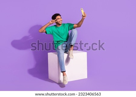 Full size cadre of young cheerful blogging guy shooting selfie cadre sitting white box podium new video isolated on violet color background