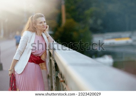 pensive elegant woman in pink dress and white jacket in the city on the bridge. Royalty-Free Stock Photo #2323420029
