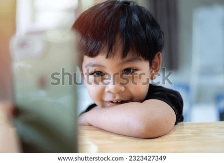 Portrait of cute little Asian resting at wooden table at home. Choosing favorite music or cartoons,  internet, watching video, playing games on smartphone.