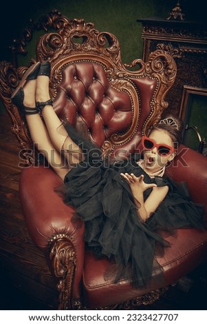 Evening Fashion for girls. A pretty little girl dressed in a puffy black dress and with elegant hairstyle and jewelry blows kisses to fans. An expensive classic interior.