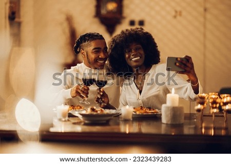 Romantic african american couple dating at night at home in a cozy atmosphere with red wine and candlelight while taking selfie with a smartphone. Happy black couple celebrating anniversary.