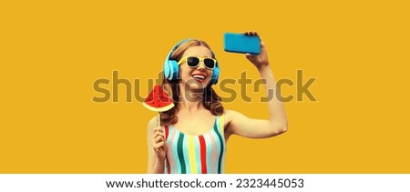 Summer portrait of cheerful happy laughing young woman taking selfie with smartphone in headphones listening to music with juicy lollipop or ice cream shaped slice of watermelon on yellow background