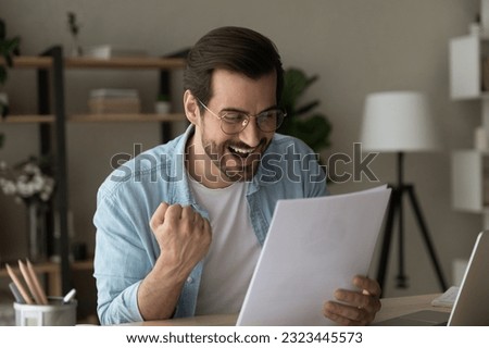 Overjoyed young man in eyeglasses looking through paper document, feeling excited of reading amazing news in letter celebrating getting lottery win notification or bank mortgage loan approval. Royalty-Free Stock Photo #2323445573