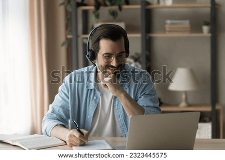Happy young man in eyewear and wireless headset with microphone looking at computer screen, listening educational lecture studying on online courses, improving knowledge distantly, writing notes. Royalty-Free Stock Photo #2323445575