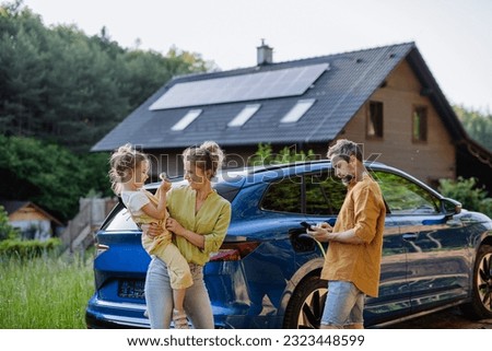 Family with little girl standing in front of their house with solar panels on the roof, having electric car. Royalty-Free Stock Photo #2323448599