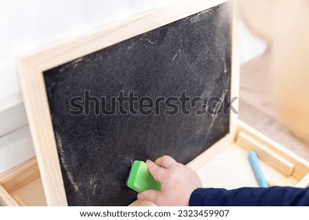 A close up portrait of a toddler drawing, scribbling or doodling a bit on a chalkboard. The child is wiping the blackboard and has a green sweeper in its hand.