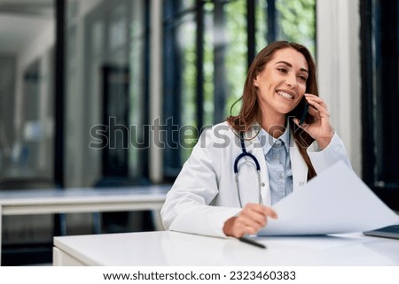 A smiling brunette female doctor talking on a phone while holding a document at the ambulance table. Royalty-Free Stock Photo #2323460383