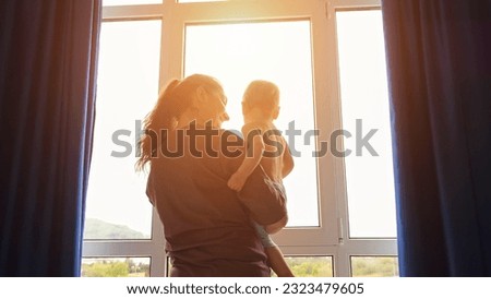 Adult woman with toddler girl in arms opens curtains together on panoramic windows. Mother and daughter look at morning cityscape from home, sunlight