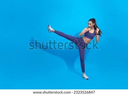 Asian female athlete workout, practice leg kicks, kicking air in sportswear. Muscular trained woman kicking with raised feet, exercise kickboxing moves, blue background. Royalty-Free Stock Photo #2323526847