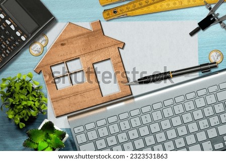 Small wooden model house on a desk with blank sheet of paper with copy space, calculator, folding ruler, drawing compass, Euro coins, pencil and a computer keyboard.
