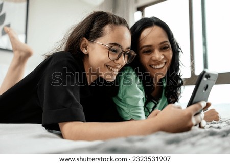 Mother and teenage daughter using smartphone together lying in bed at home. Latina mother and daughter looking at phone watching social media videos while relaxing in bedroom. family and technology Royalty-Free Stock Photo #2323531907