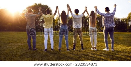 Diverse friends meet in the park. Group of multiracial young people standing in row on green lawn and holding hands up in evening sunlight. Back view, from behind. Friendship, community concept