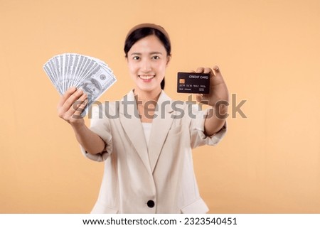 portrait of successful happy confident young asian business woman wearing white jacket holding cash money dollars and credit card standing over beige background. millionaire business, shopping concept