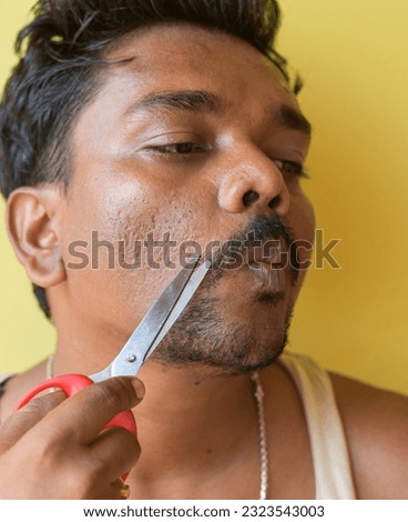 Image of an Indian young man, trimming his beard with scissors on yellow clean background at home. Selective focus. Vertical photo.