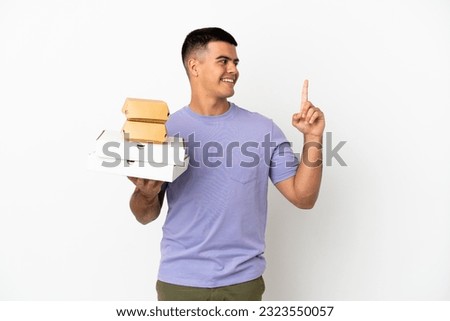Young handsome man holding pizzas and burgers over isolated white background pointing up a great idea