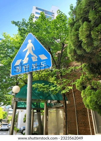 Blue and white road sign against a bus stop and trees, translation: pedestrian crossing