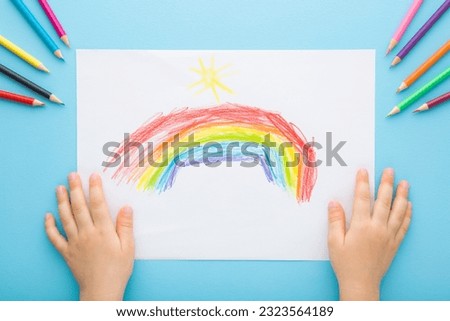 Little child hands showing colorful rainbow and sun shapes on white paper. Color pencils on light blue table background. Pastel color. Closeup. Point of view shot. Children creativity time.