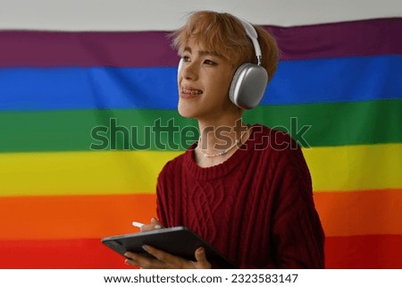 Portrait image of Young LGBT teenage boy happily smiling while wearing headphones and using tablet, pride flag on the background.