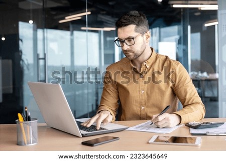 Serious young man accountant, auditor, analyst works in the office at the table behind the laptop and with documents. Writes invoices, makes analysis, fills out financial reporting and taxation. Royalty-Free Stock Photo #2323645649