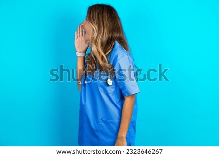 Half-faced close up portrait of astonished amazed young beautiful doctor woman standing over blue studio background holding hand near mouth