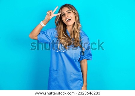 young beautiful doctor woman standing over blue studio background Doing peace symbol with fingers over face, smiling cheerful showing victory