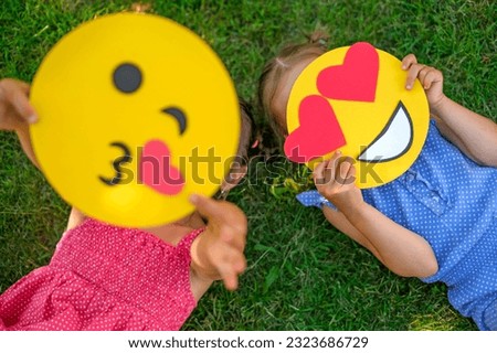 A child shows a kissing emoticon lying on the lawn grass in close-up. The girl hides behind a smiley face in love