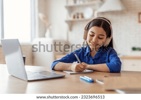 Online Education. Cheerful Arabic Little Girl In Headphones Studying With Laptop And Taking Notes, Writing Her School Homework At Desk At Home Interior. Kid Schoolgirl Learning Remotely