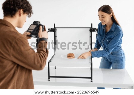 Team of male photographer and his female assistant doing content photoshoot for fast food restaurant, taking photos of burger on white platform, woman helping while working in team in photostudio