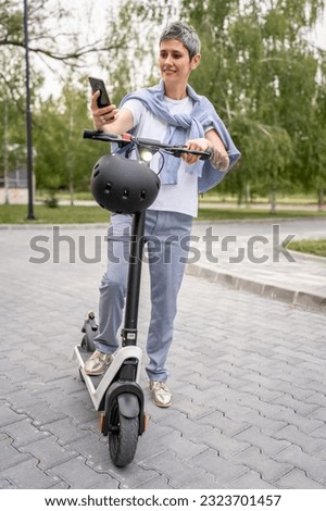 one woman mature senior caucasian female standing outdoor use mobile phone for selfie photos or video call by electric kick scooter in day confident modern alternative mode of transport copy space Royalty-Free Stock Photo #2323701457