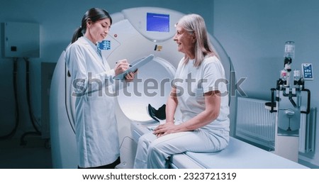 Remote shooting. Female doctor is standing near patient and explaining procedure. Female patient is smiling and listening to doctor carefully. Medical worker is holding tablet and talk to patient.