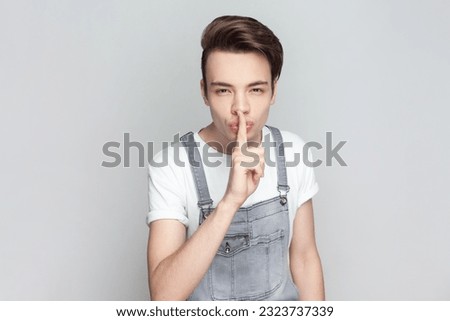 Portrait of mysterious brunette man standing presses index finger to lips, makes hush gesture tells secret, asks to be quiet, wearing denim overalls. Indoor studio shot isolated on gray background.