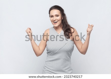 Emotional brunette woman raises clenched fists, exclaims with excitement, rejoices sweet success, feels taste of victory, wearing striped dress. Indoor studio shot isolated on gray background. Royalty-Free Stock Photo #2323740217