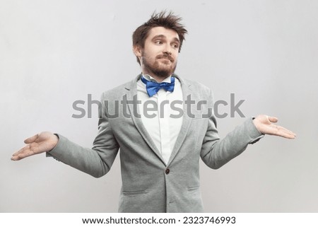 Portrait of uncertain handsome bearded man spreads hands, shrugging shoulders, doesn't know how to find solution, wearing grey suit and blue bow tie. Indoor studio shot isolated on gray background. Royalty-Free Stock Photo #2323746993