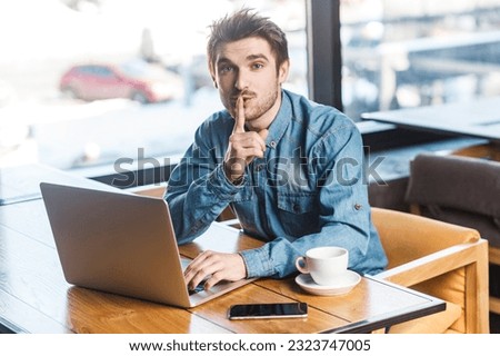 Portrait of handsome bearded young man freelancer in blue jeans shirt working on laptop, showing shh gesture, looking at camera, asking not to make noise. Indoor shot near big window, cafe background.