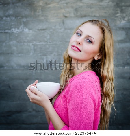 Happy young blond woman drinking hot tea or coffee and looking puzzled over gray brick wall copy space background