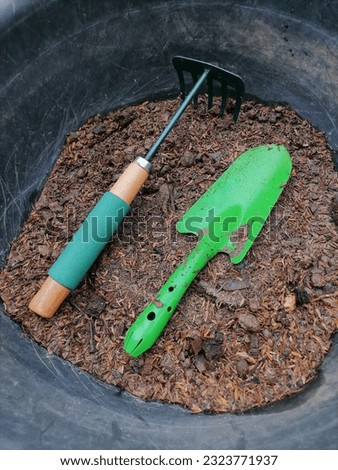 Green gardening tools are shovels and rakes placed in the soil pan. The background image is trees, leaves, lawns. Royalty-Free Stock Photo #2323771937