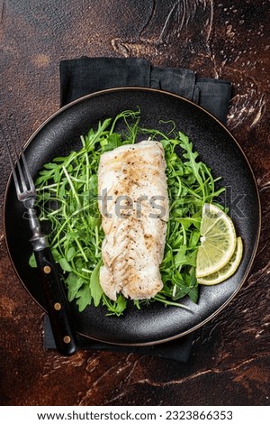 Grilled cod fish fillet served with green salad in a plate. Dark background. Top view. Royalty-Free Stock Photo #2323866353