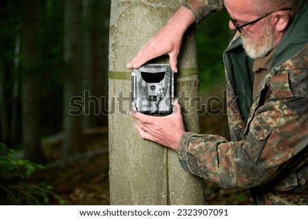 Hunter sets a trail camera on a tree in the forest. Trail cameras are often used by hunters for automatic photography or video shooting of wildlife in the forest.