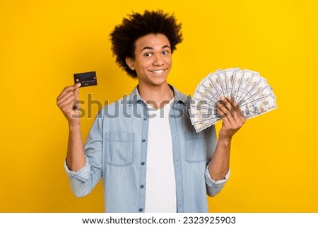 Photo of cute positive guy dressed denim shirt comparing bank card cash fan isolated yellow color background