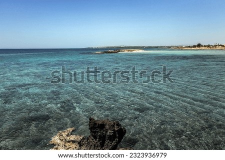 The azure water of the Mediterranean Sea in the bay of Nissaki Agias Theklas with black rocks against the background of an island with a sandy shore under a blue sky (Sotira, Ayia Napa, Cyprus)