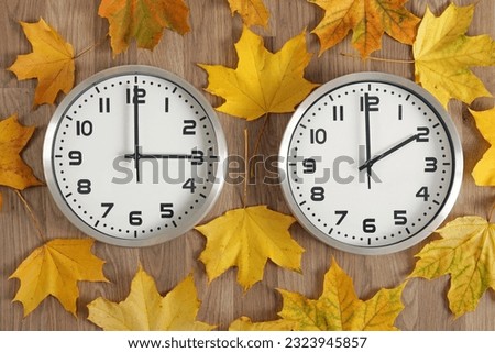 Two clocks, one shows three o'clock, the other shows two o'clock. Yellow fallen autumn leaves lie around. Symbol of time change from summer time to winter time. Moving the hands backwards. Royalty-Free Stock Photo #2323945857