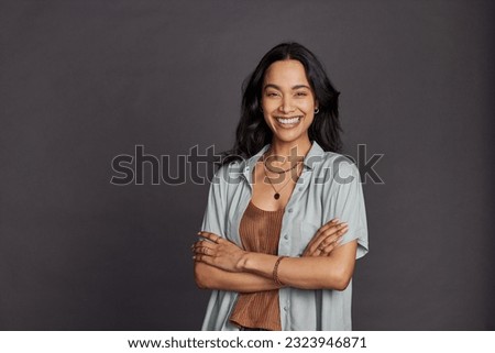 Portrait of smiling young multiethnic woman in casual clothing with arms crossed standing against grey background. Cheerful latin woman with folded hands looking at camera with big grin.  Royalty-Free Stock Photo #2323946871