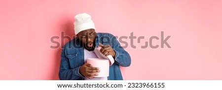 Image of surprised african-american man open box with birthday gift, staring at present with amazement, standing against pink background.